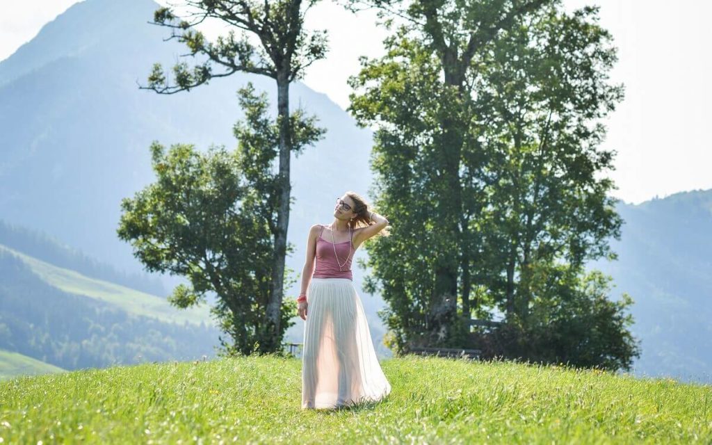 Epigenetische Wirkung von Ayurveda Eine Frau in einem rosa Oberteil und weißen langen Rock steht auf einer Wiese, im Hintergrund steht ein Baum