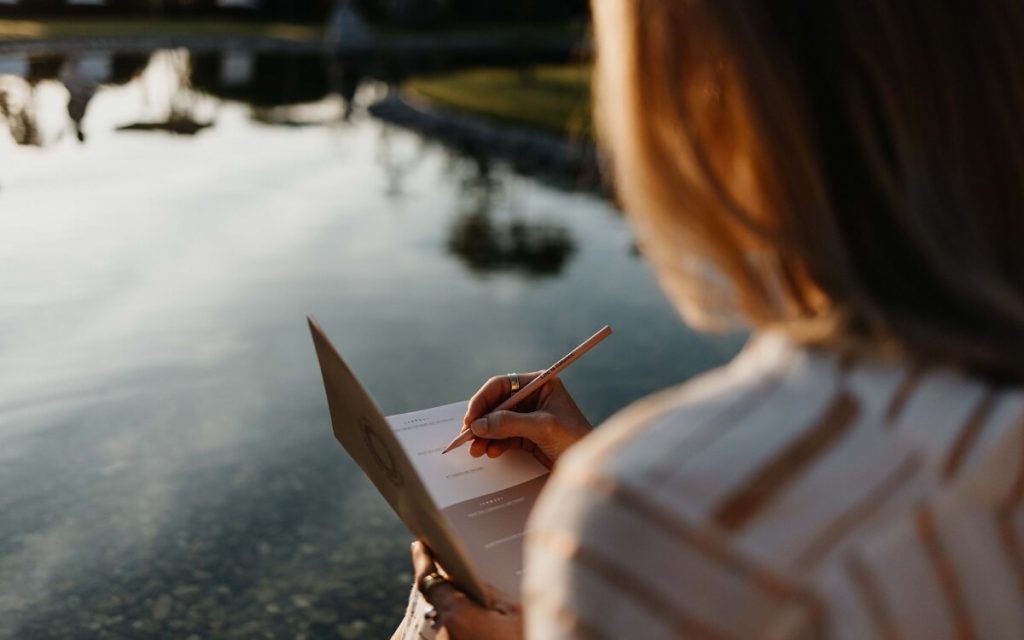 Mind Detox für Longevity Eine Frau sitzt am Wasser und schreibt in einem Journal; Mind Detox für Longevity