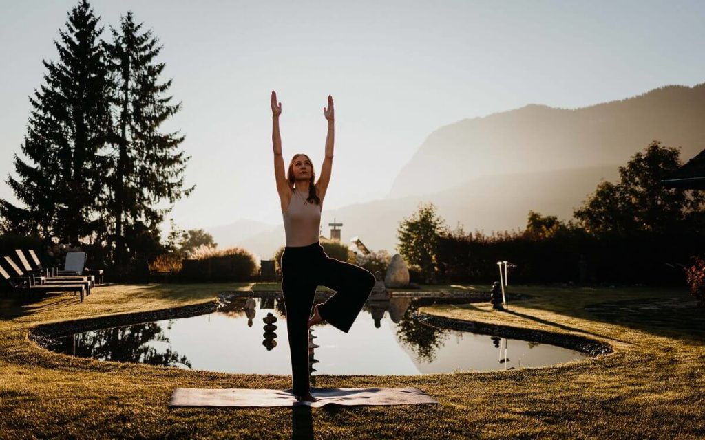 Rituale für Longevity im European Ayurveda Eine Frau steht in der Baum-Pose vor einem Teich im Morgengrauen; Rituale für Longevity im European Ayurveda
